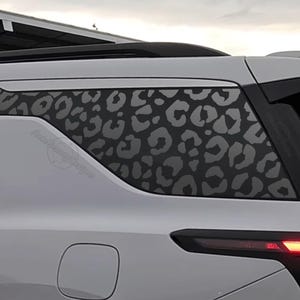 May include: White SUV with a black leopard print design on the rear side window. The design is a repeating pattern of gray spots. The vehicle has a black roof rack and a red taillight.