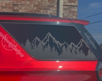 May include: A red car with a black window decal of a mountain range with trees. The decal says "Auto Beauty Designs" on the side.