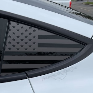 May include: A white car with a black American flag decal on the rear window. The decal is in the shape of a triangle and features a black and white American flag design. The text "Auto Beauty Designs" is visible in the lower right corner of the image.