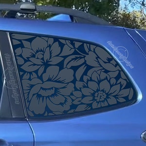 May include: A blue car with a floral design on the back window. The design is a dark gray color and features large flowers with intricate details. The text "Auto Beauty Designs" is visible on the window.