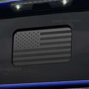 May include: A black truck tailgate with a gray American flag decal. The flag is in the center of the tailgate and is surrounded by a black border.