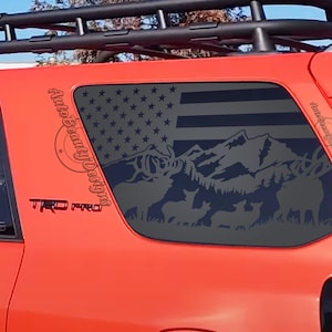 May include: A red car with a black roof rack and a black and white American flag decal on the back window. The decal also features a mountain scene with a deer and a fawn. The car has a "TRD PRO" emblem on the side.