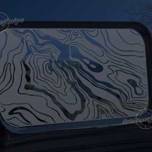 May include: A black and white topographic map design decal on a car window. The decal is a gray color with black lines. The decal is on the back window of a car. The decal is a popular design for outdoor enthusiasts.