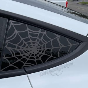 May include: A car window with a spiderweb design. The web is a light gray color and covers the entire window. The car is white, and the window has a black border. The text "AutoBeautyDesigns" is visible.