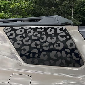 May include: A vehicle's rear window featuring a black and gray leopard print design. The window is part of a tan-colored SUV, with a black roof rack visible above. The design adds a stylish, animal-inspired aesthetic.