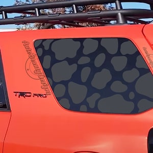 May include: A red Toyota 4Runner with a black roof rack and a black and gray camouflage pattern on the rear window. The car has a "TRD PRO" emblem on the side. The window decal says "Auto Beauty Designs".