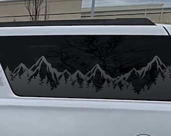 May include: A black and white mountain range silhouette decal with a forest of trees at the base. The decal is applied to the back window of a white SUV.