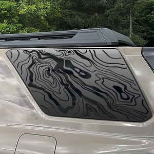 May include: A vehicle's rear window features a topographic map design in black. The window is tinted, and the vehicle's body is a light metallic color. The roof rack and spoiler are black, and the background shows green trees.
