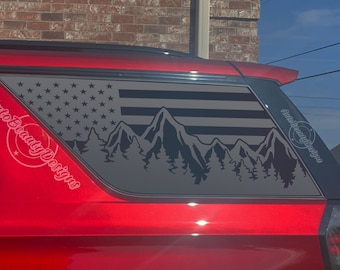 May include: A red car with a black and gray American flag and mountain silhouette decal on the rear window. The decal has the text "Auto Beauty Designs" on the side.