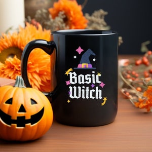 May include: Black ceramic mug with a white graphic of a witch's hat and the text "Basic Witch". The mug is surrounded by autumnal decor, including pumpkins, sunflowers, and autumn leaves.