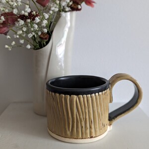 Peut inclure: Une tasse en céramique avec un motif texturé en bois dans des tons de brun et beige. La tasse a une bordure et une poignée noires.