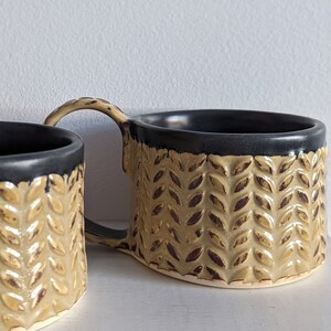 May include: Two ceramic mugs with a black rim and a gold leaf pattern. The mugs are handmade and have a rustic look.