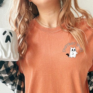 May include: A burnt orange long-sleeved t-shirt with a white ghost graphic and the text "Everybody say Boo!" in black. The shirt is being worn by a person with a black and white plaid shirt layered over it.