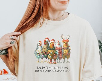 May include: Ivory unisex t-shirt with a Christmas-themed design featuring five hens wearing festive attire and the text "Holidays with the hens the ultimate cluster flock".