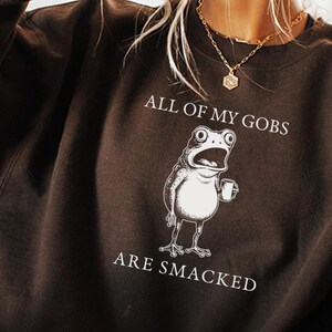 Puede incluir: Sudadera de color chocolate oscuro con el texto "ALL OF MY GOBS ARE SMACKED" y un dibujo de una rana con una taza. La sudadera es de cuello redondo y manga larga.