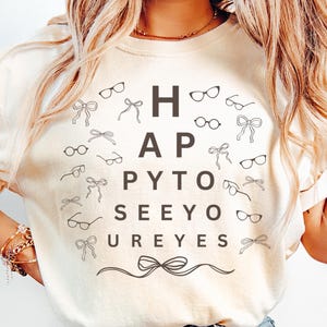 May include: Ivory-colored t-shirt with the text "HAPPY TO SEE YOUR EYES" in a playful eye chart design. The shirt features eyeglasses and bow accents. The brand "Comfort Colors" is visible at the bottom.