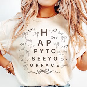 May include: Ivory-colored t-shirt with the text "HAPPY TO SEEYO URFACE" in black, surrounded by eyeglasses and bow tie graphics. The shirt is from Comfort Colors.