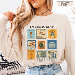 Pode incluir: Camiseta de manga comprida cor marfim com um gr&aacute;fico semelhante a um selo. O gr&aacute;fico inclui o texto "IN MEMORIAM" e v&aacute;rios selos com o ano "2025" e frases como "Public Broadcasting", "Vaccine Research" e "USAID Global Aid".
