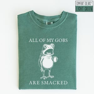 Puede incluir: Camiseta verde claro Comfort Colors con el texto "ALL OF MY GOBS ARE SMACKED" y una ilustraci&oacute;n blanca de una rana sosteniendo una taza. La camiseta es de color liso y tiene cuello redondo.