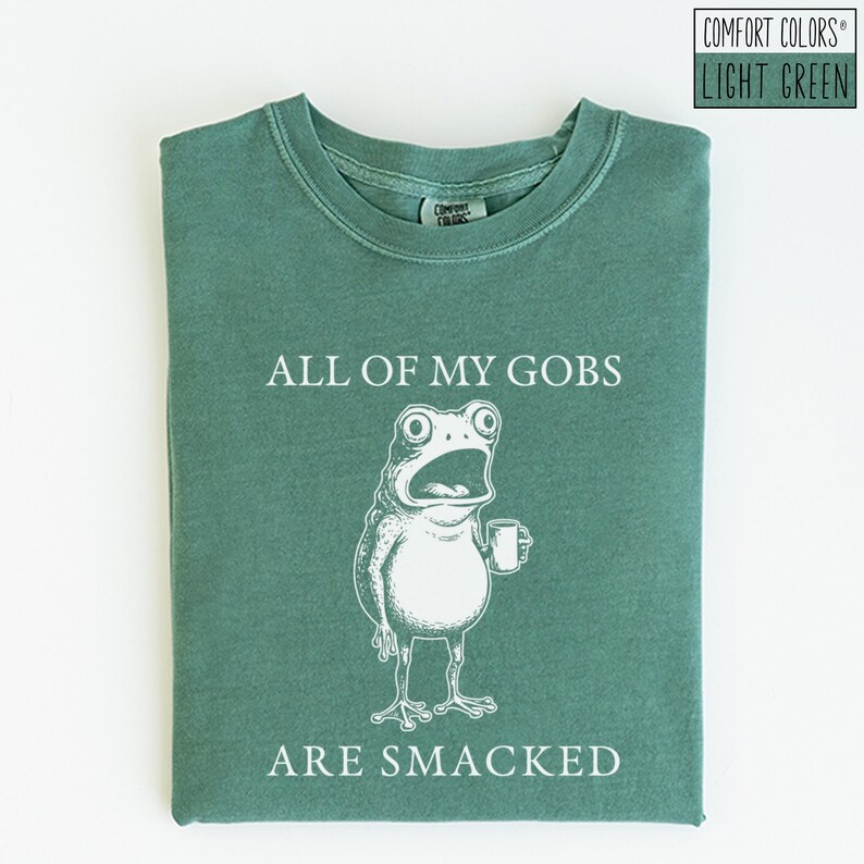 Puede incluir: Camiseta verde claro Comfort Colors con el texto "ALL OF MY GOBS ARE SMACKED" y una ilustraci&oacute;n blanca de una rana sosteniendo una taza. La camiseta es de color liso y tiene cuello redondo.