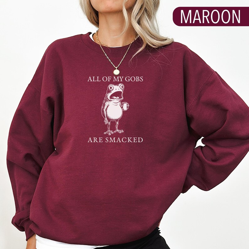 Puede incluir: Sudadera burdeos con el texto "ALL OF MY GOBS ARE SMACKED" y una ilustraci&oacute;n de una rana de dibujos animados con una taza. La sudadera es de color burdeos liso. El texto y la ilustraci&oacute;n de la rana son blancos.