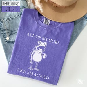 Puede incluir: Una camiseta violeta Comfort Colors con el texto "ALL OF MY GOBS ARE SMACKED" y una rana de dibujos animados sosteniendo una taza. La camiseta est&aacute; doblada sobre unos vaqueros azules, un collar y un sombrero marr&oacute;n.