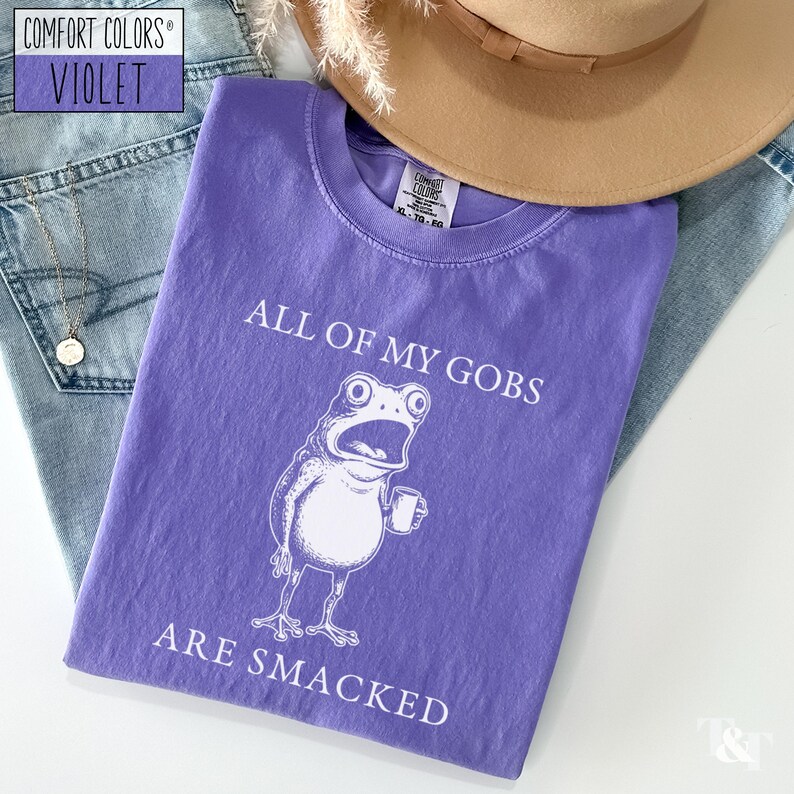 Puede incluir: Una camiseta violeta Comfort Colors con el texto "ALL OF MY GOBS ARE SMACKED" y una rana de dibujos animados sosteniendo una taza. La camiseta est&aacute; doblada sobre unos vaqueros azules, un collar y un sombrero marr&oacute;n.