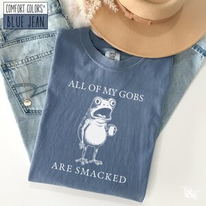 Puede incluir: Una camiseta azul Comfort Colors con el texto "ALL OF MY GOBS ARE SMACKED" y una rana de dibujos animados sosteniendo una taza. La camiseta est&aacute; colocada sobre unos vaqueros azules y un sombrero beige.