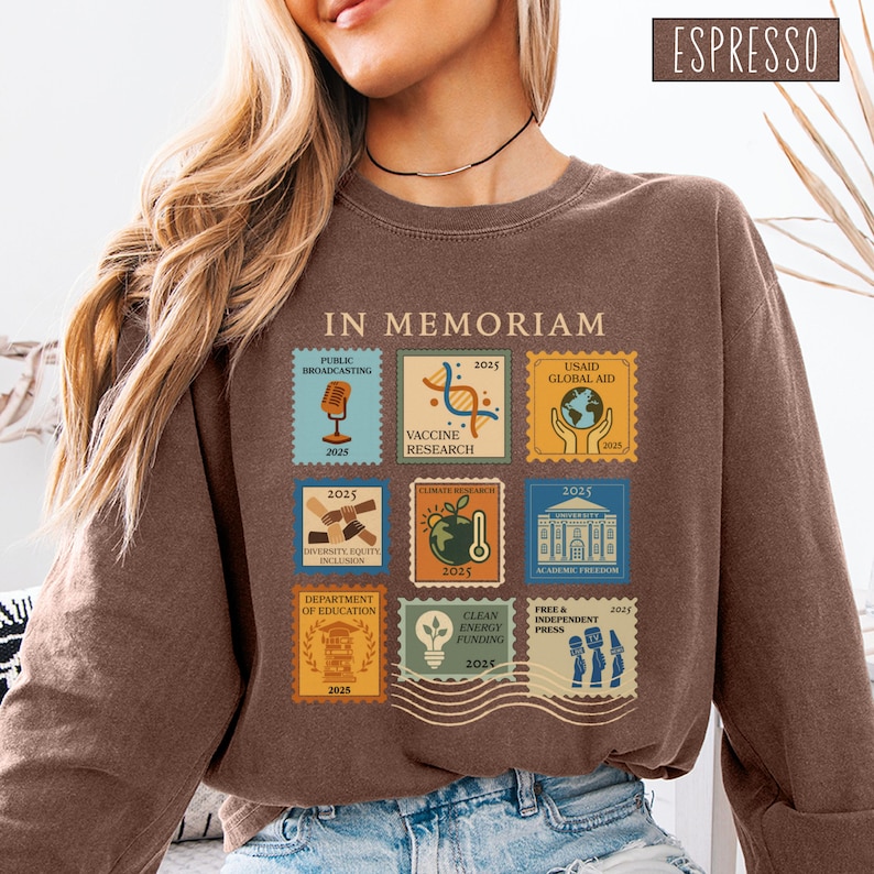 Pode incluir: Camisola cor espresso com um gr&aacute;fico de selos postais. Os selos apresentam ilustra&ccedil;&otilde;es e texto, incluindo "IN MEMORIAM", "Public Broadcasting", "Vaccine Research", "USAID Global Aid", "Diversity, Equity, Inclusion", "Climate Research", "Academic Freedom", "Department of Education", "Clean Energy Funding" e "Free & Independent Press".