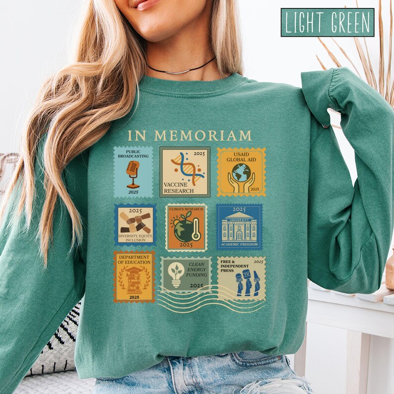 Pode incluir: Camisola verde claro com um gr&aacute;fico semelhante a um selo que diz "IN MEMORIAM". Os selos apresentam imagens e texto relacionados com a radiodifus&atilde;o p&uacute;blica, investiga&ccedil;&atilde;o de vacinas, ajuda global da USAID, diversidade, investiga&ccedil;&atilde;o clim&aacute;tica e financiamento de energia limpa.
