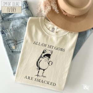 Puede incluir: Camiseta marfil Comfort Colors con un gr&aacute;fico negro de una rana sosteniendo una taza y el texto "ALL OF MY GOBS ARE SMACKED". La camiseta est&aacute; colocada sobre unos vaqueros azul claro, un collar y un sombrero marr&oacute;n.