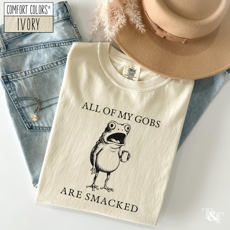 Puede incluir: Camiseta marfil Comfort Colors con un gr&aacute;fico negro de una rana sosteniendo una taza y el texto "ALL OF MY GOBS ARE SMACKED". La camiseta est&aacute; colocada sobre unos vaqueros azul claro, un collar y un sombrero marr&oacute;n.