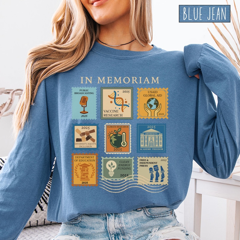 Pode incluir: Uma camisa azul jeans de manga comprida com um gr&aacute;fico de selos postais. Os selos apresentam texto e ilustra&ccedil;&otilde;es relacionadas com a radiodifus&atilde;o p&uacute;blica, a investiga&ccedil;&atilde;o de vacinas e a investiga&ccedil;&atilde;o clim&aacute;tica. A camisa tamb&eacute;m tem o texto "IN MEMORIAM".