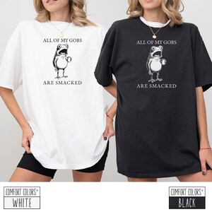 Puede incluir: Camisetas blancas y negras con el texto "ALL OF MY GOBS ARE SMACKED" y una ilustraci&oacute;n de rana de dibujos animados. Las camisetas son de la marca Comfort Colors.