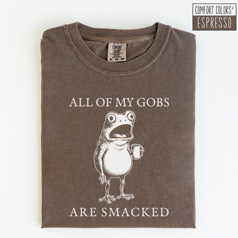 Puede incluir: Camiseta color espresso con una ilustraci&oacute;n humor&iacute;stica de una rana sosteniendo una taza. El texto "ALL OF MY GOBS ARE SMACKED" est&aacute; impreso encima y debajo de la rana. La camiseta es Comfort Colors XL-TG-EG.