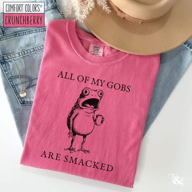 Puede incluir: Una camiseta rosa con el texto "ALL OF MY GOBS ARE SMACKED" y una ilustraci&oacute;n negra de una rana con una taza. La camiseta est&aacute; colocada sobre unos vaqueros azules y un sombrero beige.
