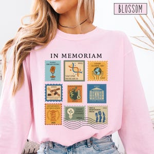 Pode incluir: Uma camisola rosa claro com a inscri&ccedil;&atilde;o "IN MEMORIAM" acima de uma grelha de nove selos postais. Cada selo apresenta um design diferente e o ano de 2025. A palavra "BLOSSOM" est&aacute; numa caixa no canto superior direito.