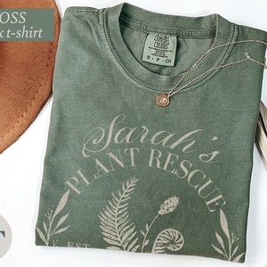 May include: A moss green unisex t-shirt with the text "Sarah's Plant Rescue Est." printed in a light brown color. The design features a fern and other botanical illustrations.