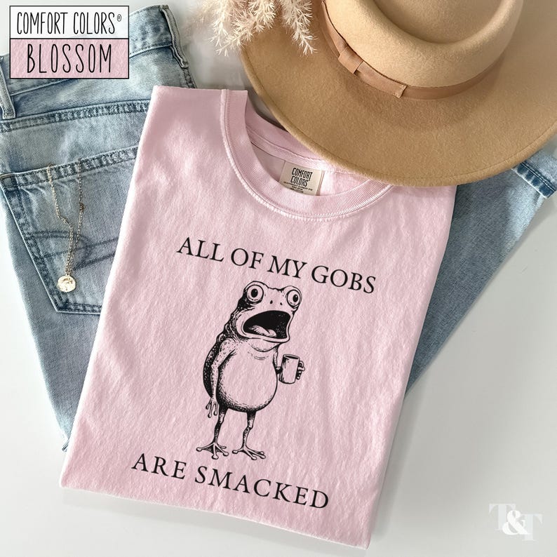 Puede incluir: Camiseta rosa claro con la frase "ALL OF MY GOBS ARE SMACKED" y una ilustraci&oacute;n en blanco y negro de una rana con una taza. La camiseta se muestra con vaqueros azules, un collar y un sombrero beige.