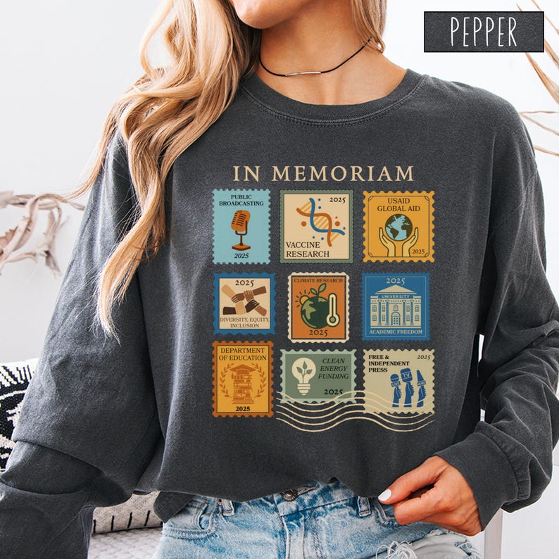 Pode incluir: Camisola cinza escuro com um design de selo. Os selos apresentam as palavras "IN MEMORIAM" e v&aacute;rios temas como "Public Broadcasting", "Vaccine Research" e "Climate Research". A palavra "PEPPER" est&aacute; no canto superior direito.
