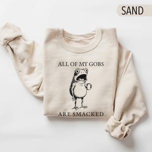 Puede incluir: Sudadera color arena con el texto "ALL OF MY GOBS ARE SMACKED" y una rana de dibujos animados con una taza. La rana tiene ojos grandes y la boca abierta. La sudadera est&aacute; doblada con las mangas enrolladas.