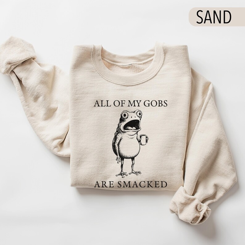 Puede incluir: Sudadera color arena con el texto "ALL OF MY GOBS ARE SMACKED" y una rana de dibujos animados con una taza. La rana tiene ojos grandes y la boca abierta. La sudadera est&aacute; doblada con las mangas enrolladas.
