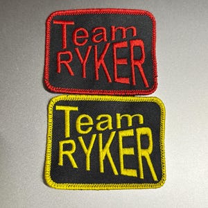May include: Two rectangular embroidered patches. The top patch has a black background with red text and border that reads "Team RYKER". The bottom patch has a black background with yellow text and border that reads "Team RYKER".