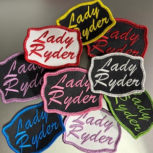 May include: Nine embroidered patches with the text "Lady Ryder" in different colors and fonts. The patches are on a gray surface.