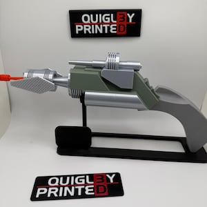 May include: A 3D-printed prop gun with a gray handle, green body, and silver accents. The gun has a red tip and is displayed on a black stand. The text "QUIGLBY PRINTED" is displayed on a black rectangle.