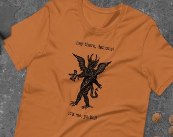 Könnte beinhalten: Ein orangefarbenes T-Shirt mit einer schwarz-weißen Illustration eines Dämons, der einen Kelch hält. Der Text auf dem Shirt lautet "hey there, demons! it's me, ya boi!"