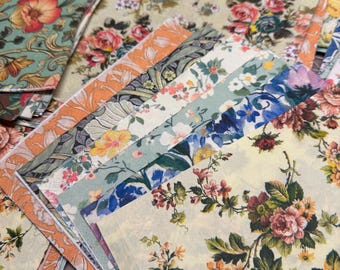 May include: A collection of square paper napkins featuring various floral patterns in shades of yellow, orange, green, blue, and red. The napkins are layered and fanned out, displaying intricate botanical designs including roses and foliage.