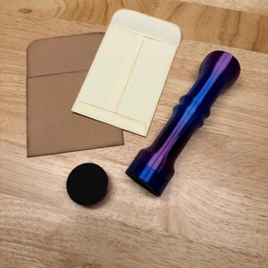 May include: A cylindrical handle with a metallic blue and purple gradient finish, featuring a textured black circular base attachment. Beside the handle are two small, rectangular paper envelopes, one in light tan and one in cream color.