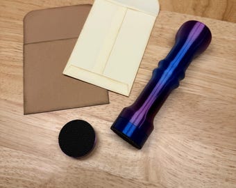 May include: A cylindrical handle with a metallic blue and purple gradient finish, featuring a textured black circular base attachment. Beside the handle are two small, rectangular paper envelopes, one in light tan and one in cream colour.