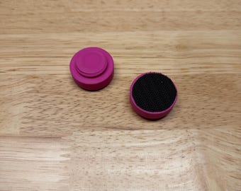 May include: Two circular controller thumbstick covers in a vibrant magenta colour. One cover displays a smooth, concave top surface, while the other features a textured black circular grip pad centred on its top surface.
